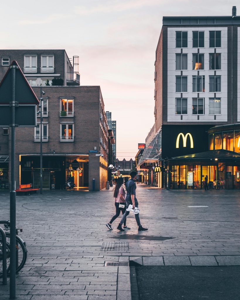 Couple walking in a city square with buildings and McDonald's at sunset.